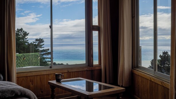 Une pièce dotée d’une grande fenêtre donnant sur l’océan et d’une terrasse verdoyante. Une tasse à café est posée sur une petite table.
