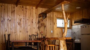 Un intérieur rustique en bois, comprenant une table à manger, des chaises, un coin cuisine et un élément décoratif original : un tronc d’arbre.