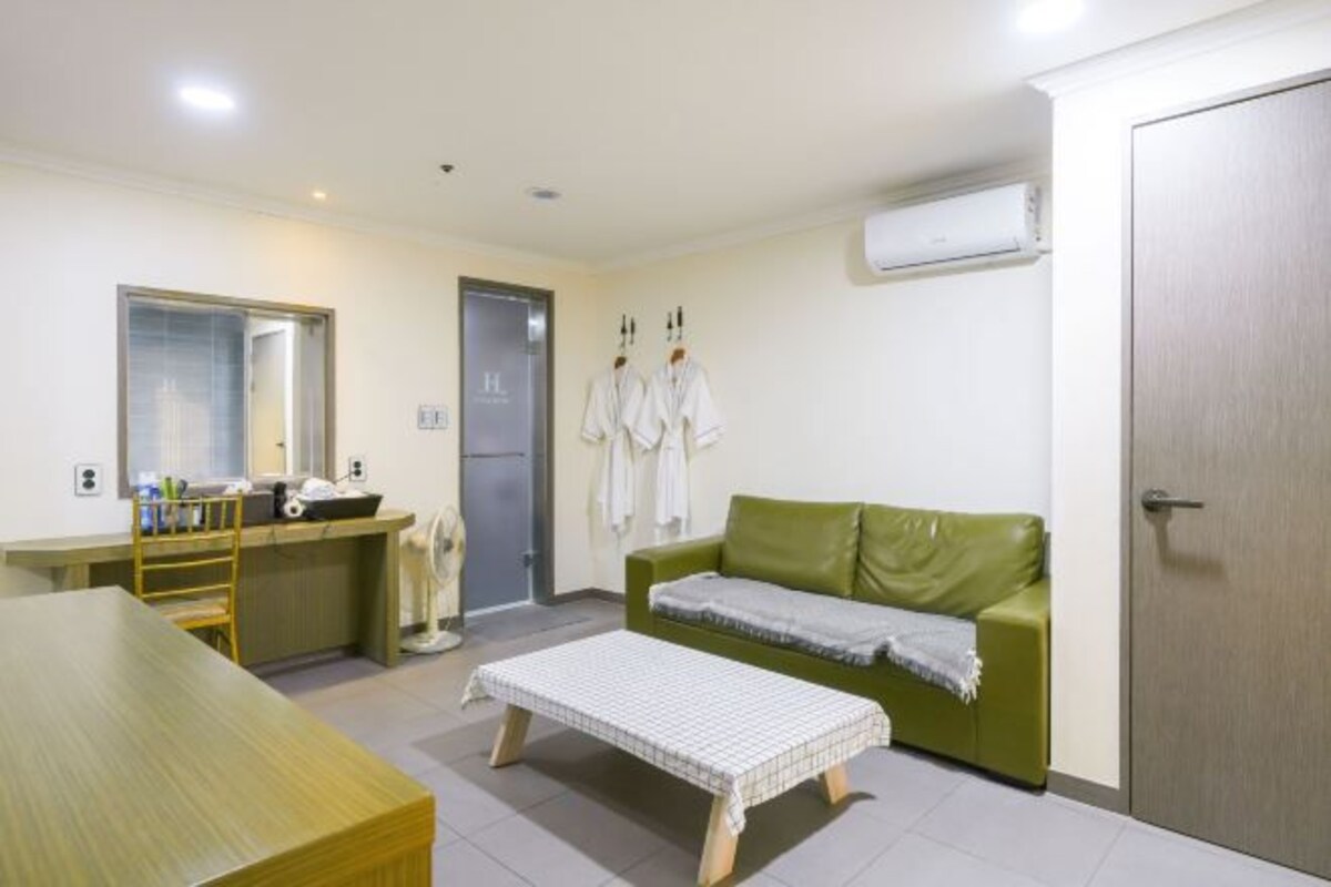A small, well-lit room with a wooden table, a green sofa, a white bench, and a mirror. There is a small kitchenette with a sink and a mirror.