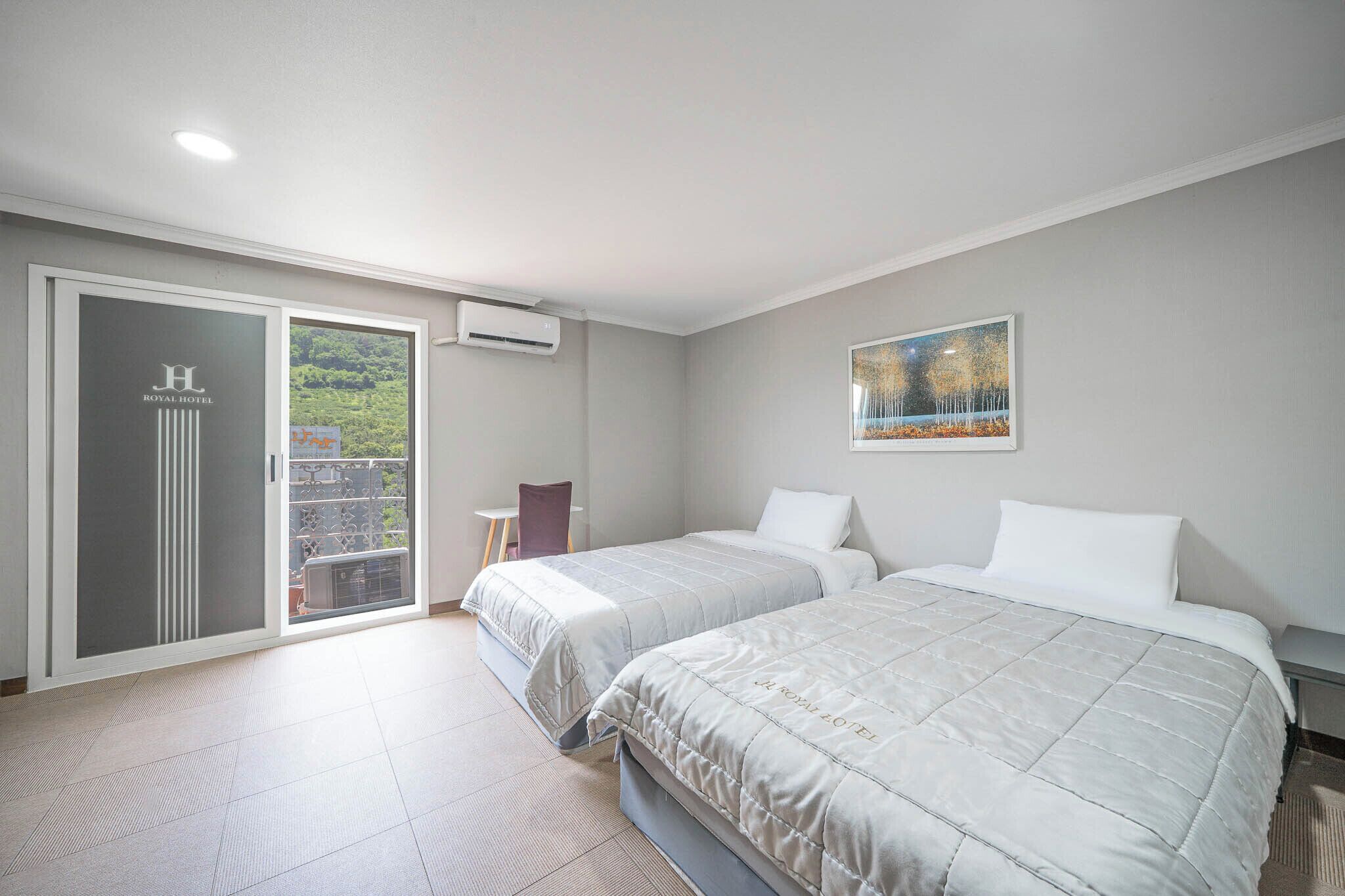 Habitación de hotel con dos camas, puerta corrediza al balcón, una mesita y un cuadro en la pared.