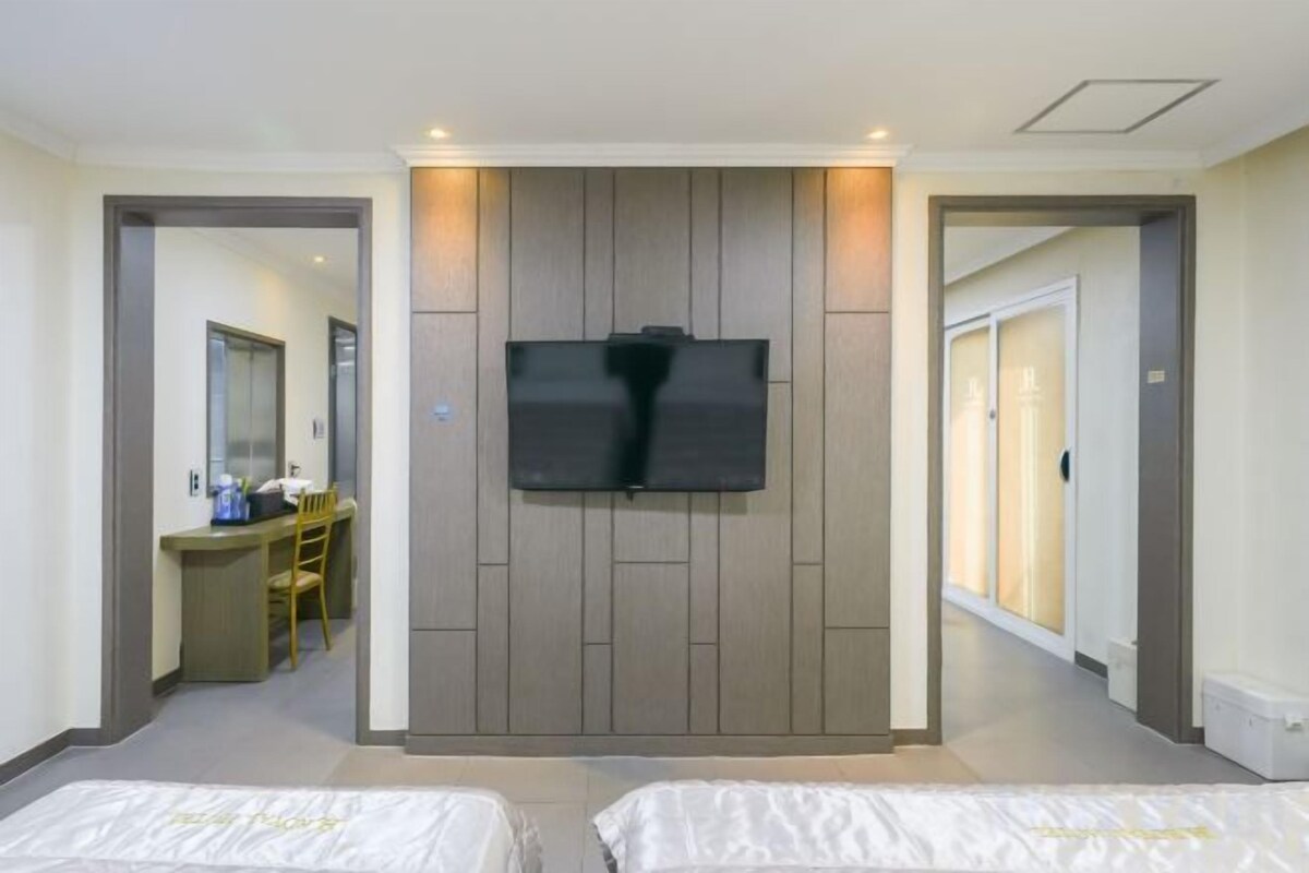 A hotel room with two beds, a TV mounted on a wooden panel wall, and a desk with a chair in the background.
