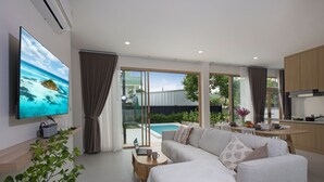 A modern living room with a large flat-screen TV, a sofa, a coffee table, and a laptop. There is a sliding glass door leading to a pool area.