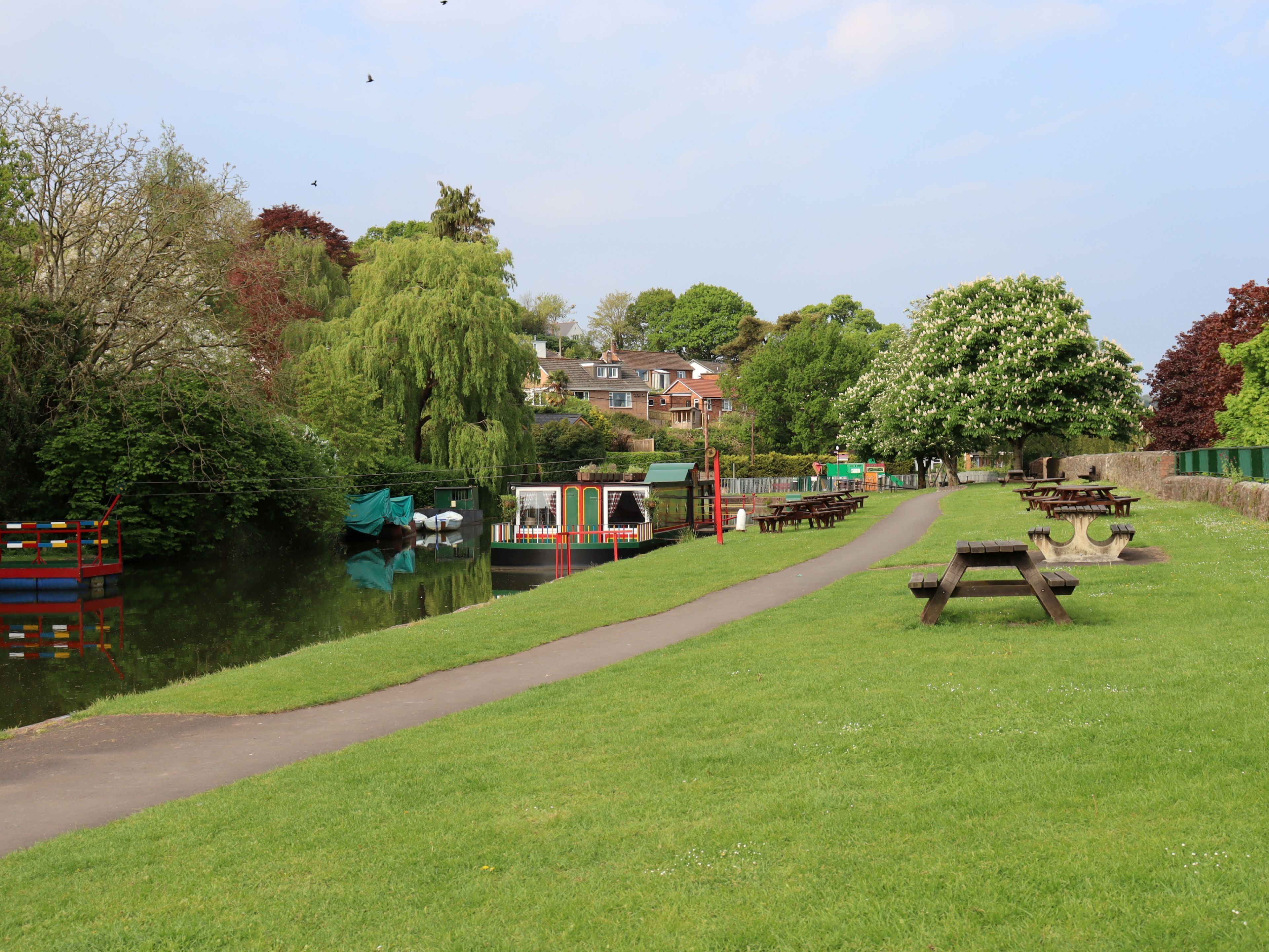 A riverside park with picnic tables, a walking path, and boats docked along the water.