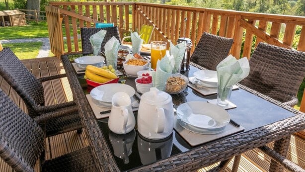 Un espace de restauration en plein air, avec des meubles en osier, une table dressée avec des assiettes, des tasses et un saladier rempli de fruits.