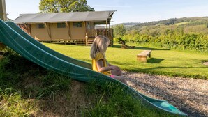 Un enfant est assis en haut d’un toboggan vert, dans un espace herbeux avec une terrasse en bois et un bâtiment en arrière-plan.