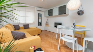 Un espace de vie moderne comprenant un canapé, une table à manger avec un plateau en verre, des chaises blanches, une guitare et une horloge murale.