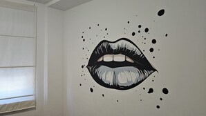Une pièce dotée d’une grande fenêtre recouverte de stores, d’un mur orné d’une fresque représentant des lèvres en noir et blanc, et d’une porte blanche.
