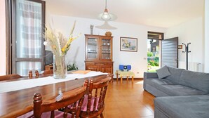 A dining area with a wooden table, chairs, a vase with dried flowers, a cabinet with glass doors, a grey sofa, and a window with a view of greenery. - Casa Lina (Colico)