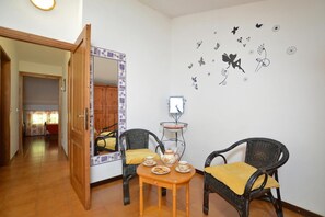 Una habitación con puerta de madera, un espejo, dos sillones de mimbre con cojines amarillos, una mesita con tetera y tazas, y una pared con adornos adhesivos.