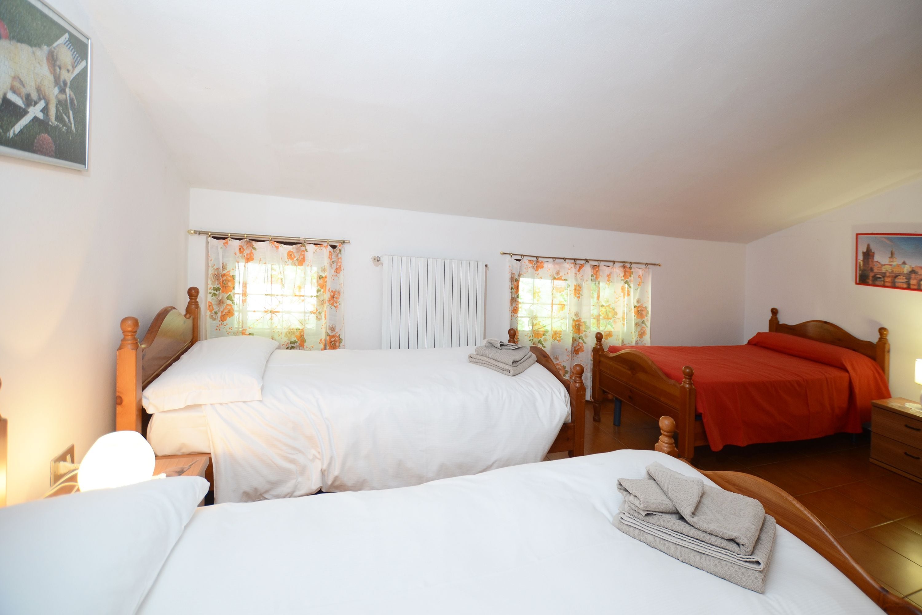 Un dormitorio con dos camas, cabeceros de madera, una ventana con cortinas florales y una mesita de noche con lámpara.