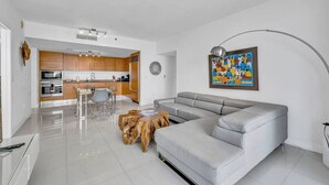 Una sala moderna con un sofá gris, una mesa de centro de madera, un comedor con sillas y una cocina al fondo.