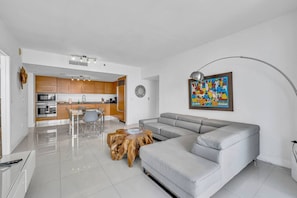 A modern living room with a grey sofa, a wooden coffee table, a dining area with chairs, and a kitchen in the background.