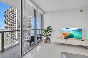 Una sala de estar moderna con un televisor grande, un balcón con vistas a edificios altos y una planta en maceta.
