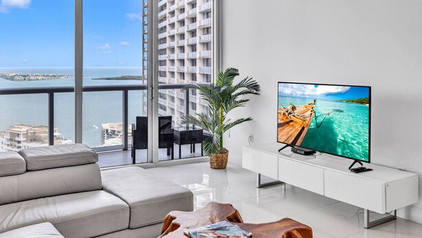 Una sala de estar moderna con un televisor grande, un sofá, una mesa de comedor y un balcón con vistas al mar y edificios.