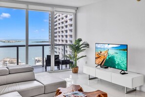Un soggiorno moderno con una grande TV, un divano, un tavolo da pranzo e un balcone vista mare e edifici.