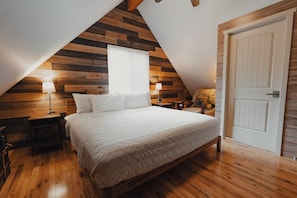 A bedroom with a sloped ceiling, wooden panel walls, a bed with a quilted bedspread, two bedside lamps, and a door.