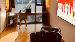 Une chambre d’hôtel moderne dotée d’une grande fenêtre donnant sur la ville, d’un bureau avec un ordinateur portable, d’une chaise et d’une valise posée sur le sol.