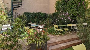 Un patio al aire libre con suelo de madera, sillas verdes y amarillas, una escalera en espiral y abundante vegetación, incluyendo plantas con flores.