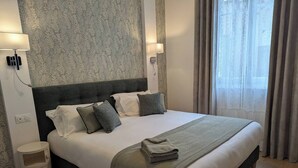 Una cama bien hecha con un cabecero gris, ropa de cama blanca y una manta gris. Se observa una mesita de noche con una lámpara y un pequeño taburete. La habitación tiene un papel pintado con motivos y una ventana con cortinas.