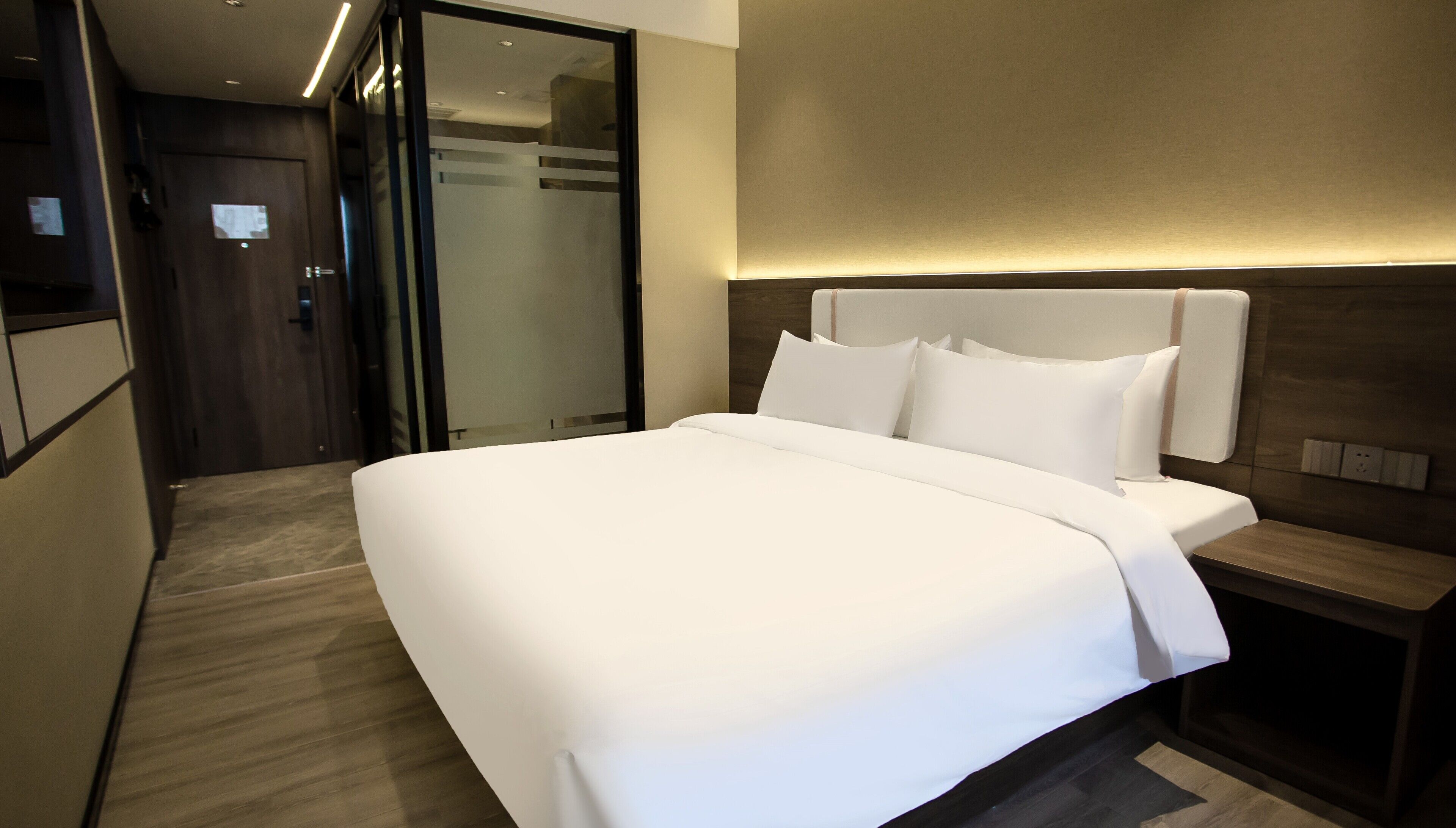 A modern hotel room with a large bed, a wooden headboard, and a glass door leading to a bathroom.