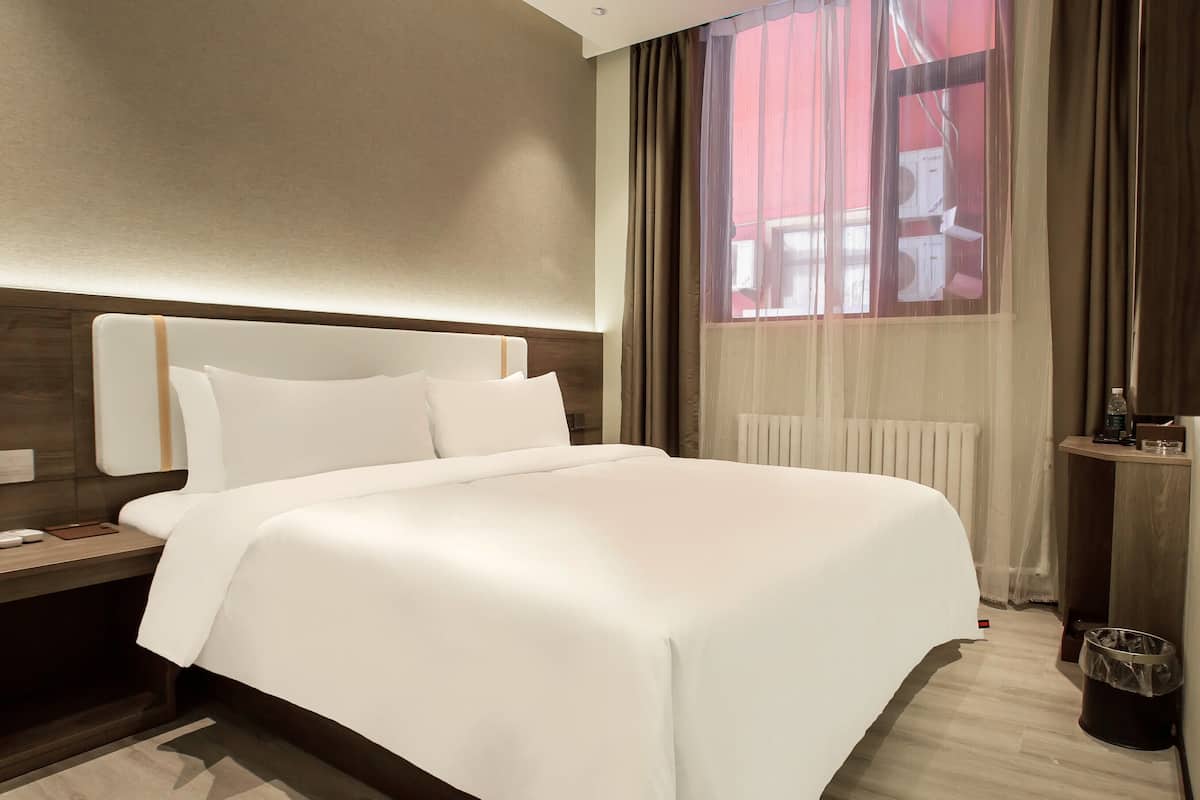 Une chambre d’hôtel avec un grand lit, une table de chevet avec une lampe, une fenêtre avec des rideaux et un radiateur.