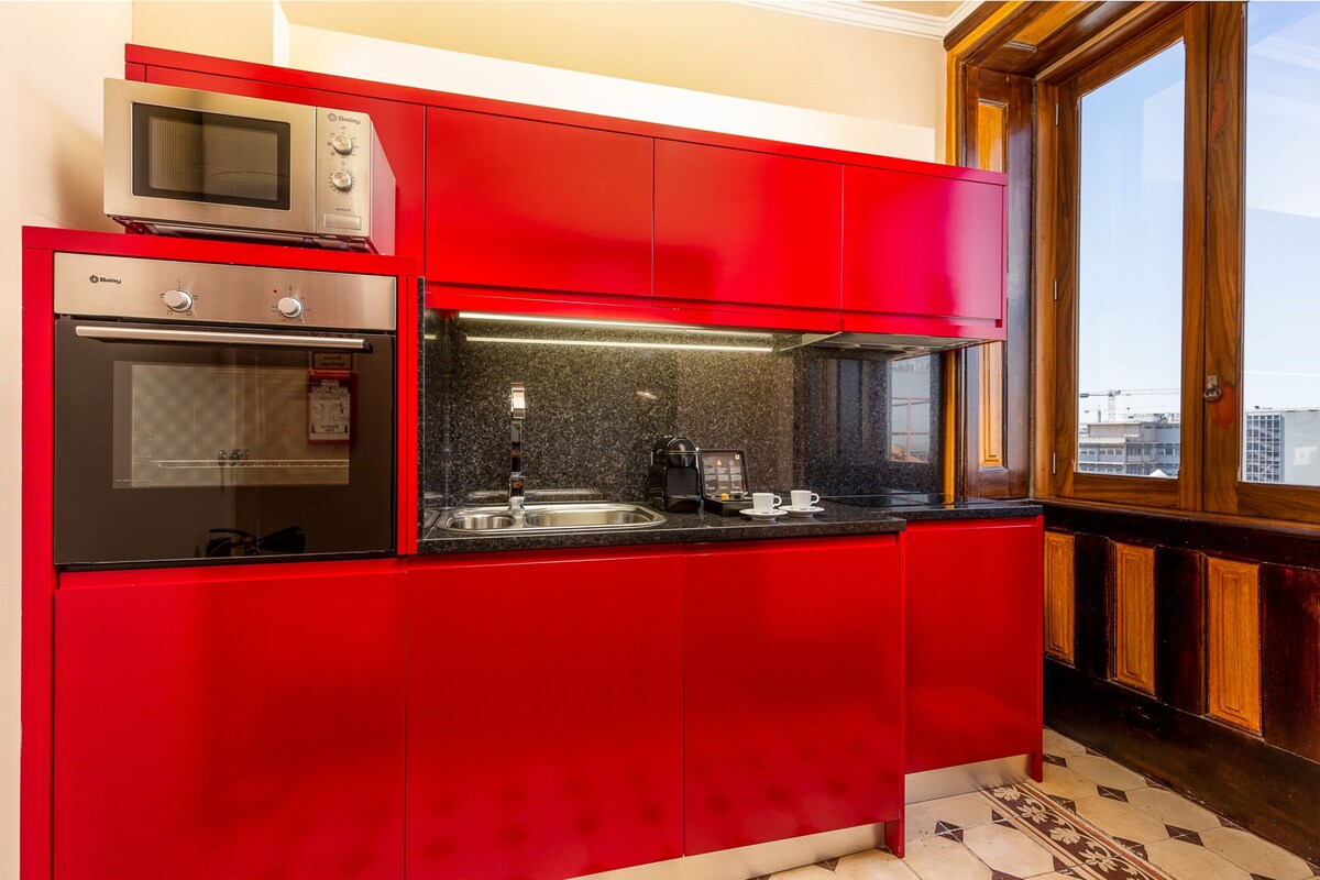 A kitchen with red cabinets, a microwave, an oven, a coffee maker, and a window with a view.