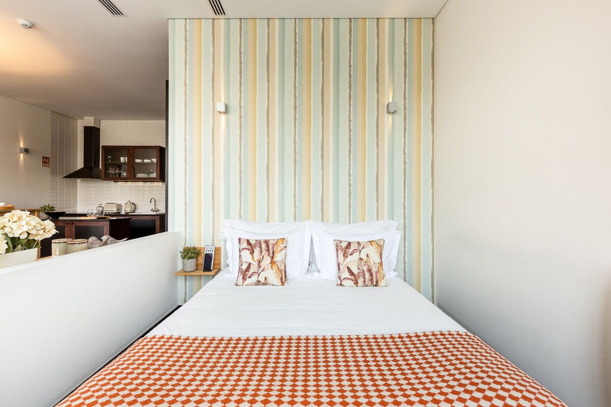 Un dormitorio con cama, papel tapiz a rayas y una cocina visible a través de una división.