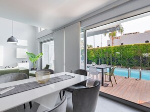 A modern dining area with a white table, grey chairs, and a large window overlooking a pool and greenery. - Miguel VIP Properties by Interhome (Albufeira)