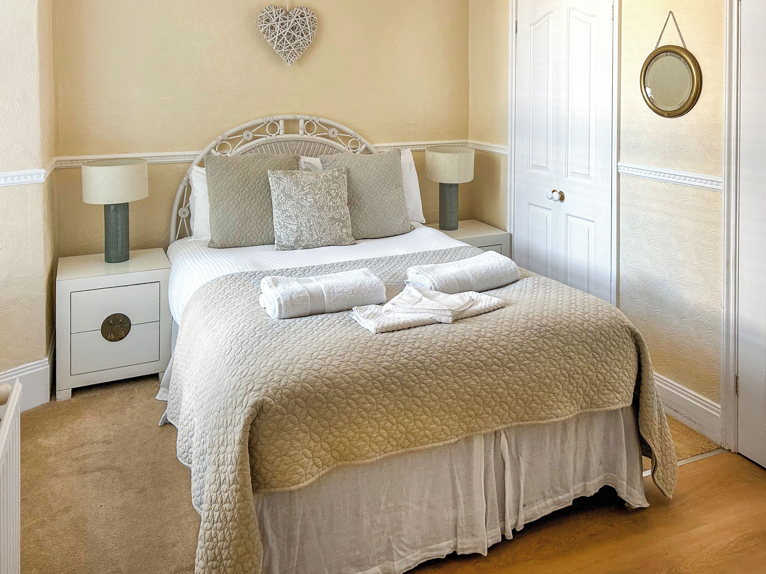 A neatly made bed with a quilted cover, two pillows, and folded towels. There is a bedside table with a lamp and a small drawer. The room has a white door and a wall-mounted heart decoration.