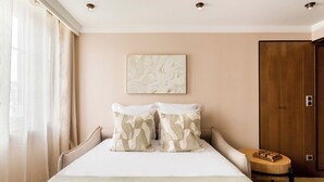 A hotel room with a large bed, a wooden wardrobe, a small round table with a wooden block, and a painting on the wall. - Merveil Luxury Suites - Bourse I-II (Paris)