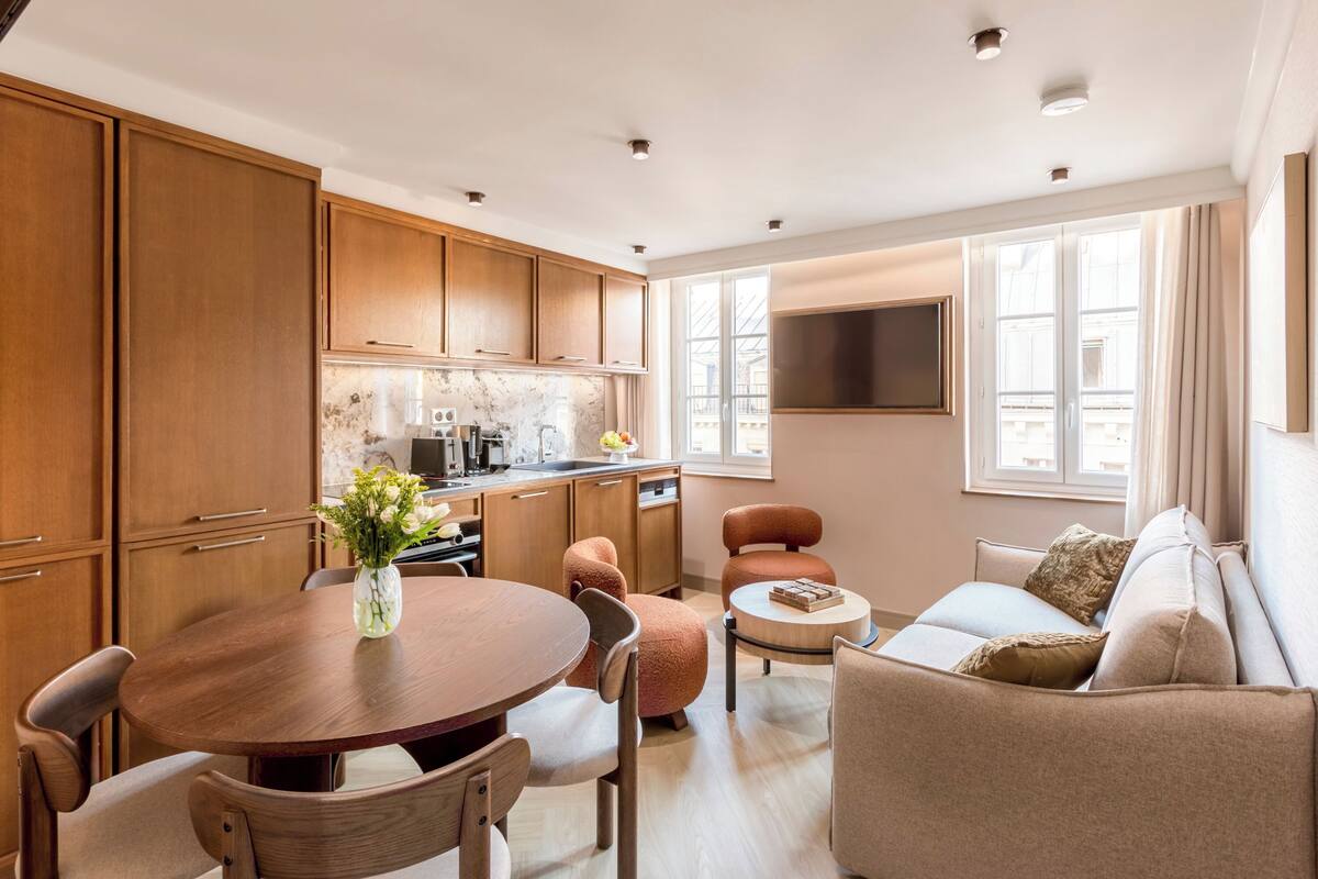A modern living room with a wooden dining table, comfortable seating, a kitchen area, and large windows providing natural light.