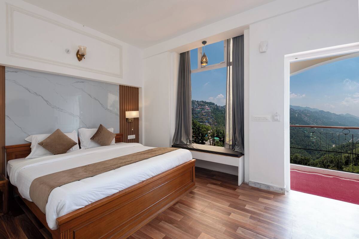 A bedroom with a large bed, a nightstand, a window with a view of a mountainous landscape, and a balcony with a red mat.