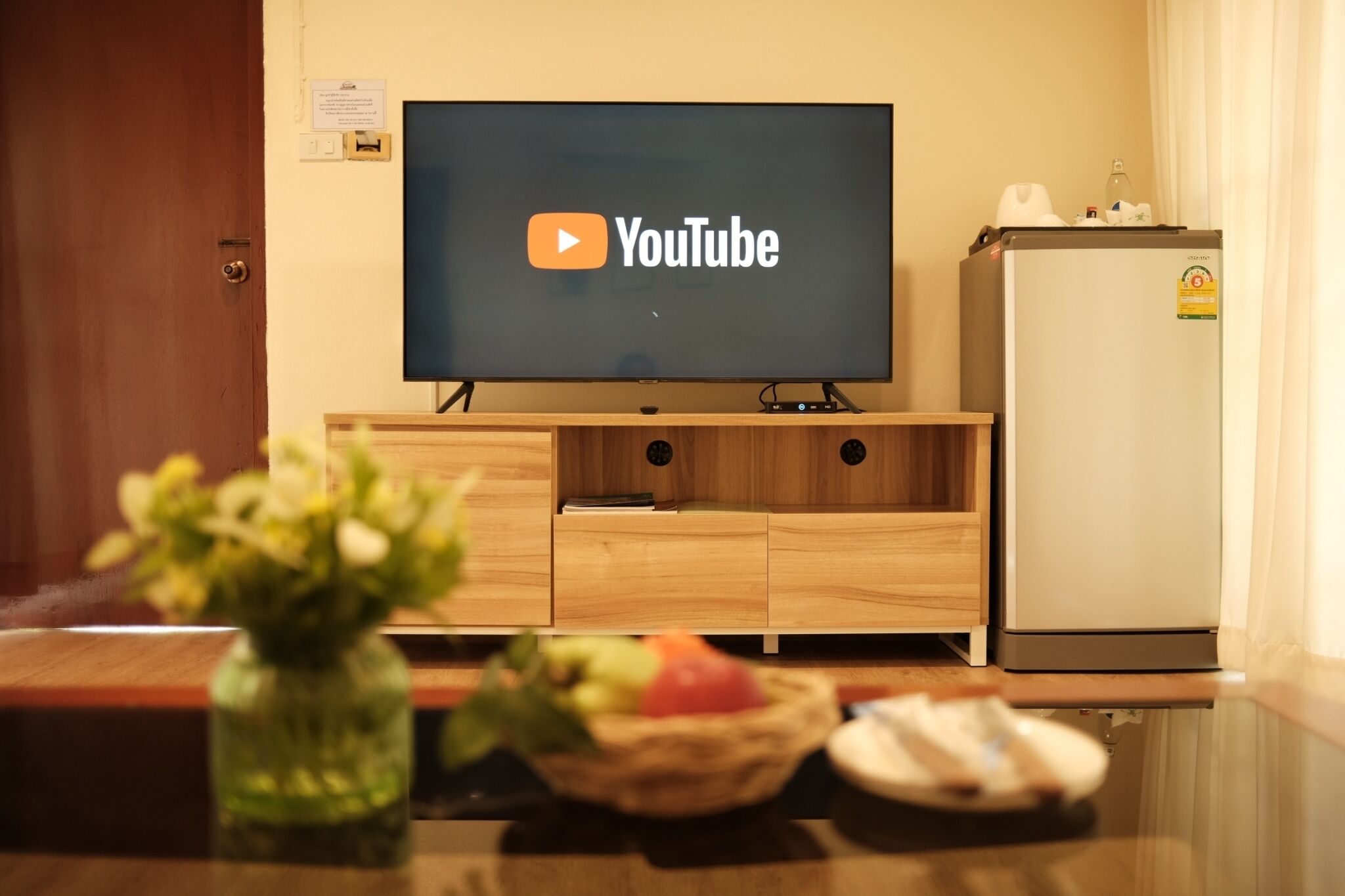 Uma televisão exibindo o logotipo do YouTube, um armário de madeira embaixo, uma geladeira e um vaso com flores sobre uma mesa.