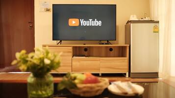 Uma televisão exibindo o logotipo do YouTube, um armário de madeira embaixo, uma geladeira e um vaso com flores sobre uma mesa.