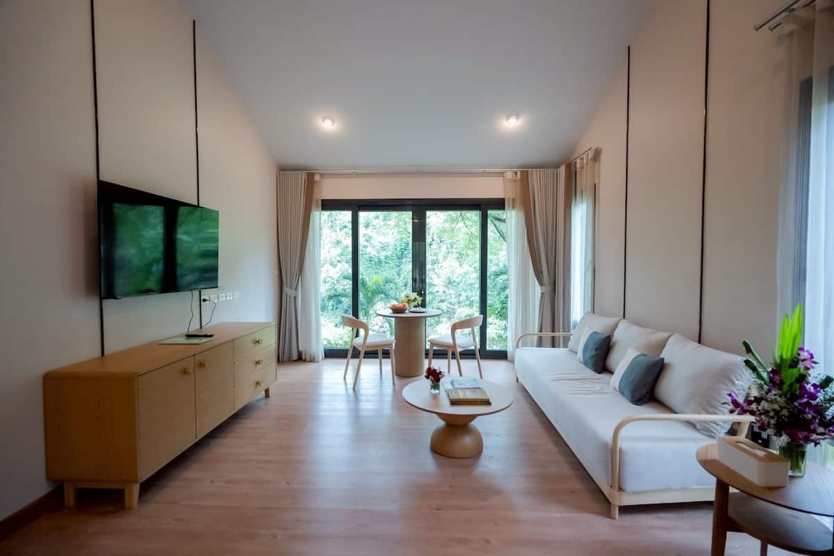 Una sala moderna con un sofá, un televisor montado en la pared, un mueble de madera, una mesa redonda de centro y vistas a la vegetación a través de grandes ventanales.