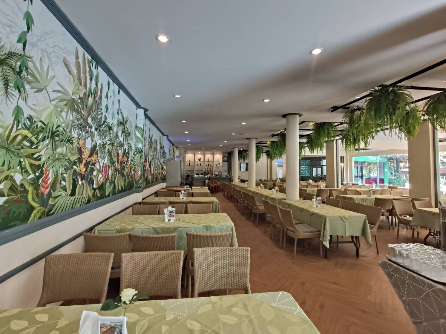 A spacious restaurant with long tables, wicker chairs, and a large mural of tropical plants on the wall.