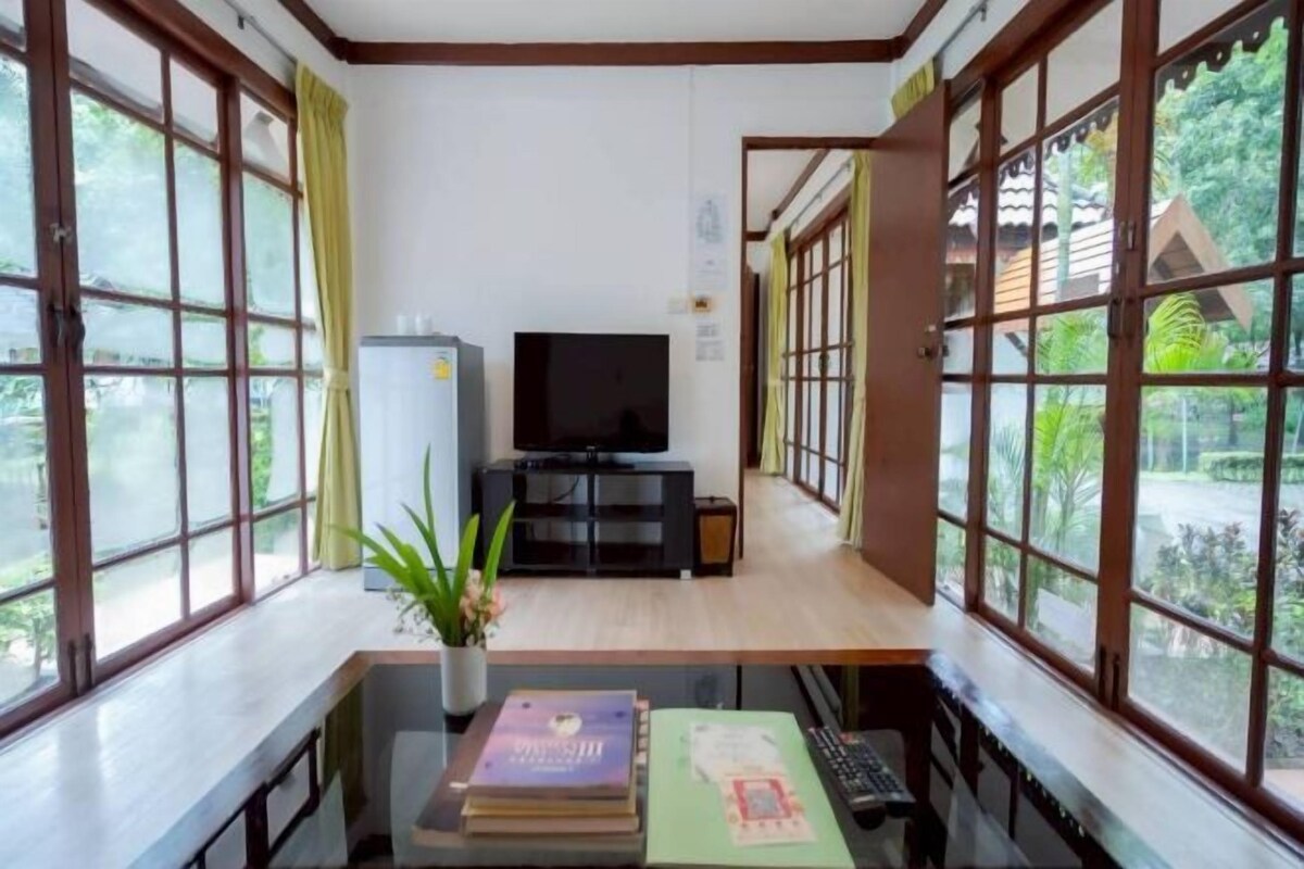 Una habitación con ventanas amplias, un televisor, una pequeña mesa con libros y una planta.