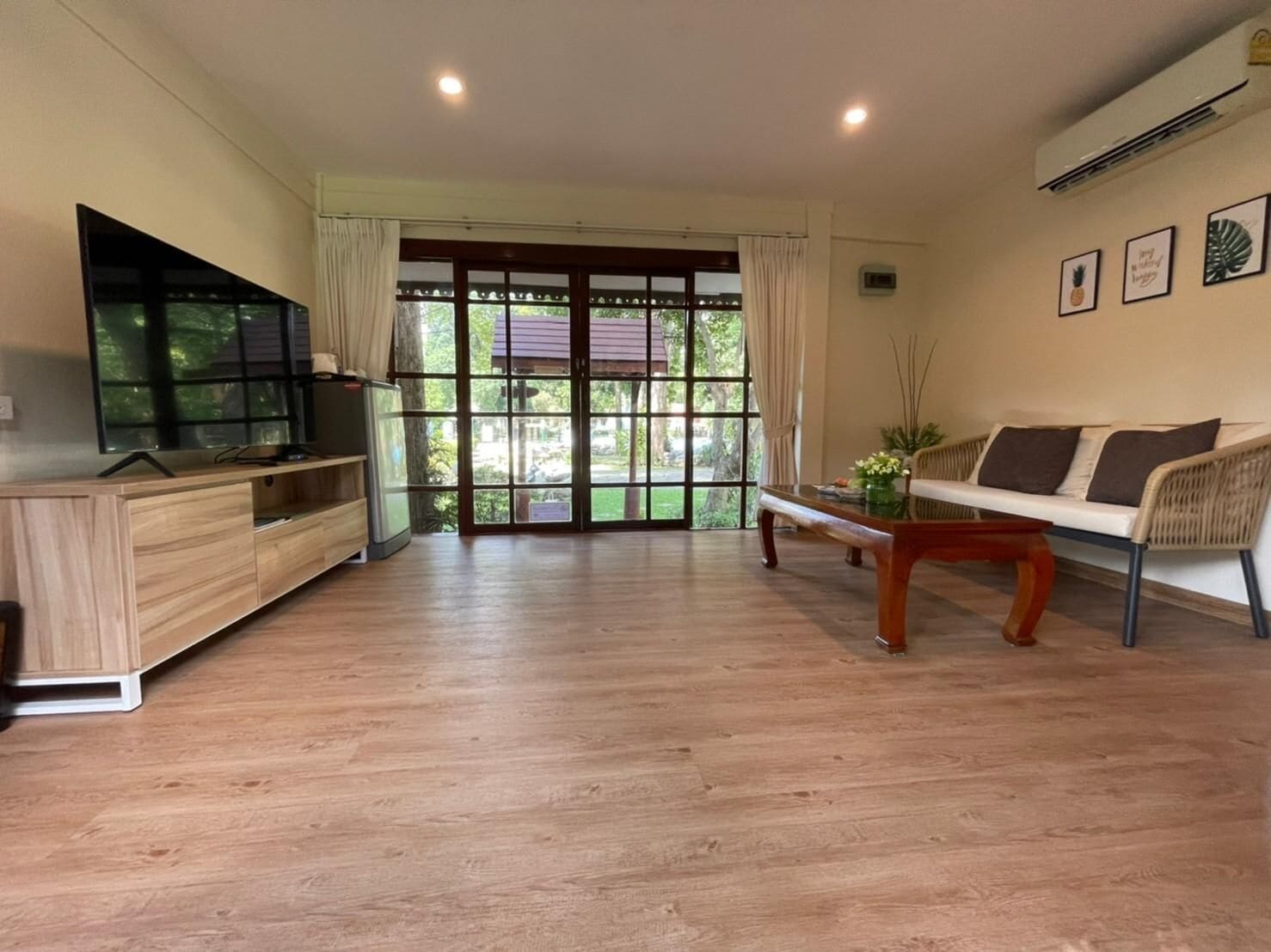 A spacious living area with a wooden floor, a sofa, a coffee table, a TV on a wooden cabinet, and large windows with a view of greenery.