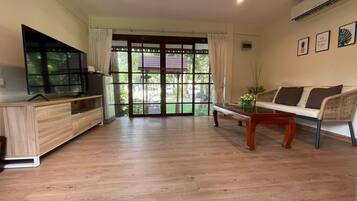 A spacious living area with a wooden floor, a sofa, a coffee table, a TV on a wooden cabinet, and large windows with a view of greenery.