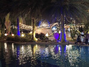 Zona junto a la piscina con luces colgantes, palmeras y una fuente de agua.