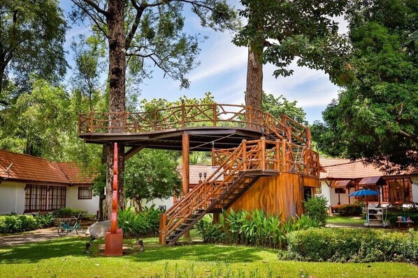 A wooden bridge connecting two buildings in a lush garden setting.