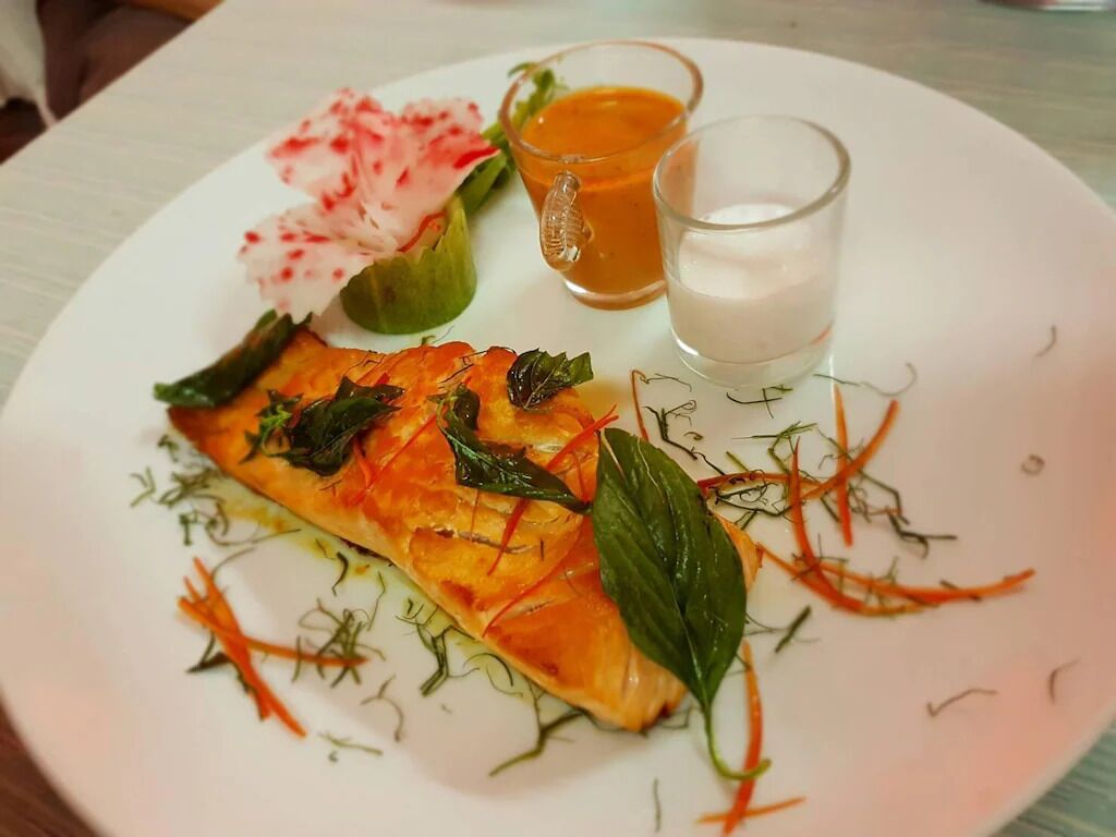 A plate with a slice of salmon tartare, garnished with herbs and thinly sliced carrots, accompanied by two glasses of sauce.
