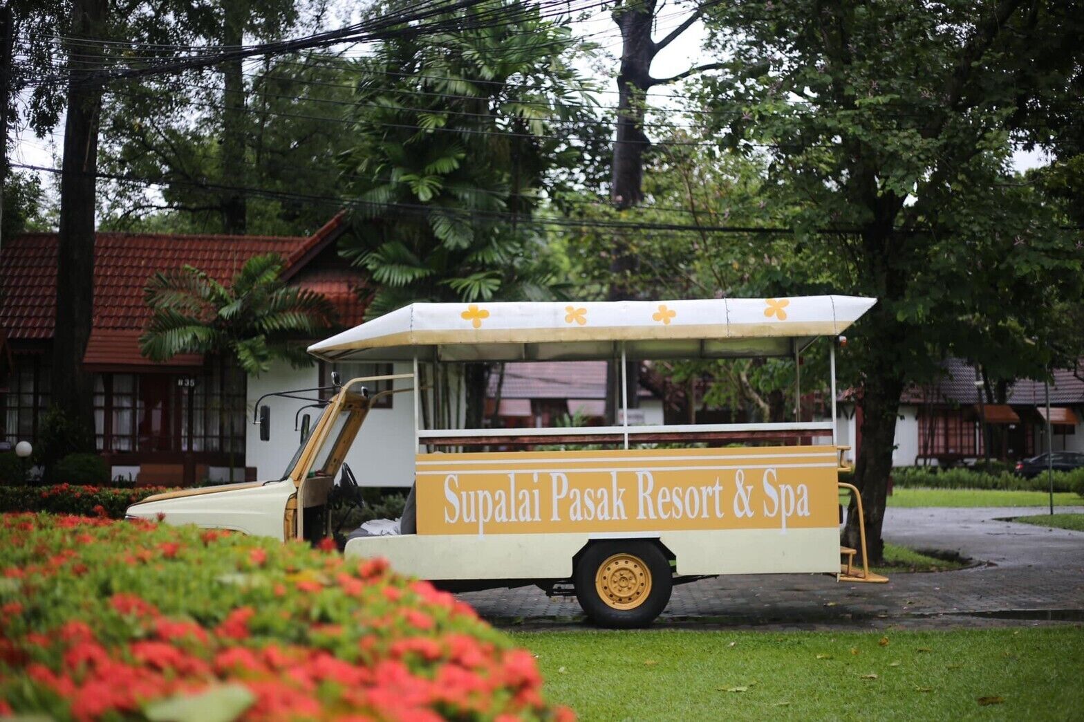 Ein weißer und gelber Food-Truck mit der Aufschrift 'Supalai Pasak Resort & Spa', der auf einer Wiese mit roten Blumen im Vordergrund geparkt ist.