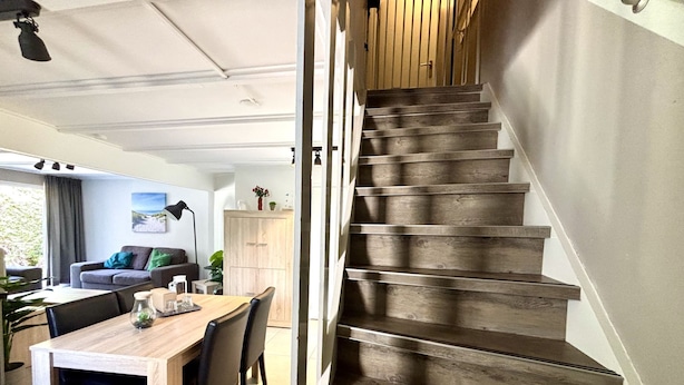 A modern interior with a staircase leading to a second floor, a dining area with a wooden table and chairs, and a living area with a sofa and a lamp.