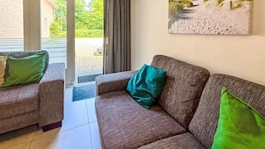 A living room with a brown sofa and two armchairs, a painting of a beach scene, and a view of a garden through a glass door.