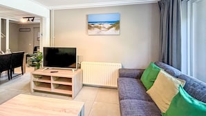 A living room with a sofa, a coffee table, a TV on a stand, a painting of a beach scene, and a window with curtains.