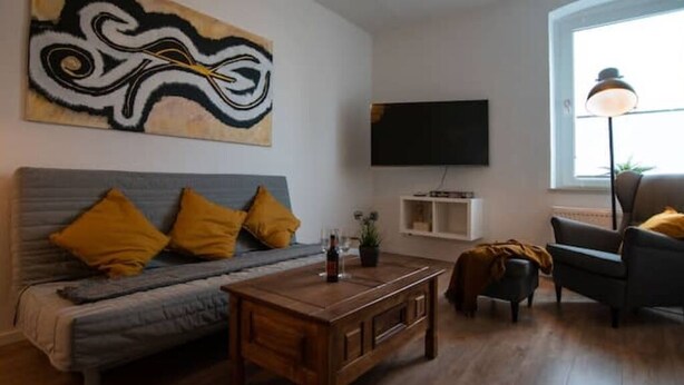 Una sala de estar con un sofá, una mesa de centro, un televisor montado en la pared y una ventana con una lámpara de pie.