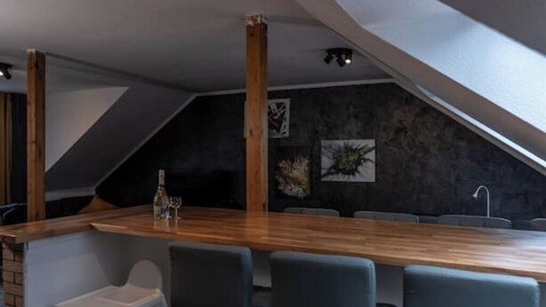 A modern bar area with a wooden counter, bar stools, and a high ceiling with exposed beams.