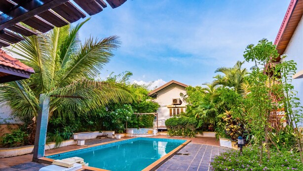 A swimming pool with a tiled deck, surrounded by lush greenery and a house in the background. - Cherry Villa,Nashik by Ebony Stays | Cosy 4BHK Villa in Nashik with Scenic Views (Nashik)