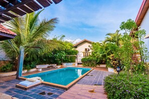 A swimming pool with a tiled deck, surrounded by lush greenery and a house in the background.